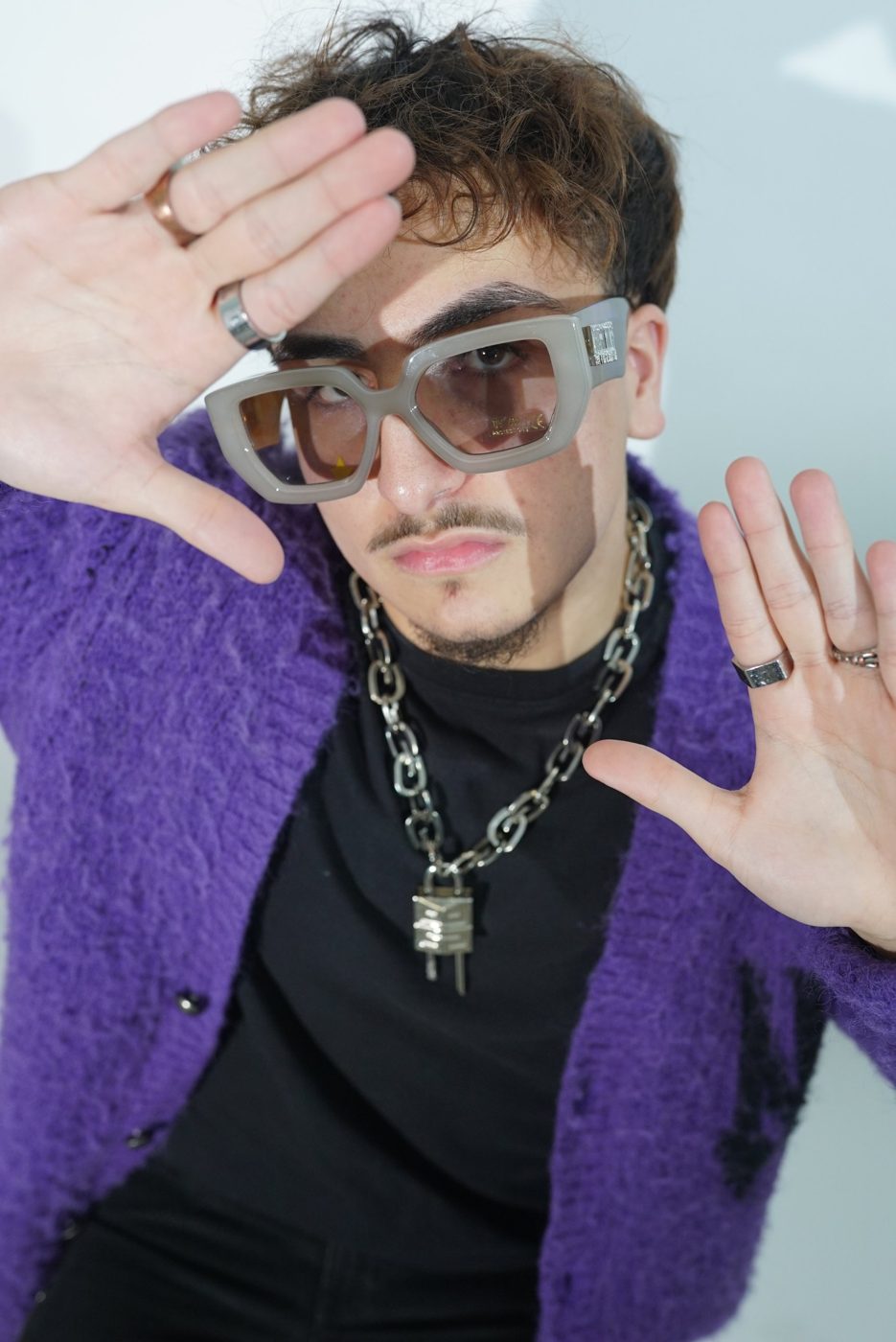 Model in a purple mohair cardigan framing his face with his hands, wearing grey statement glasses