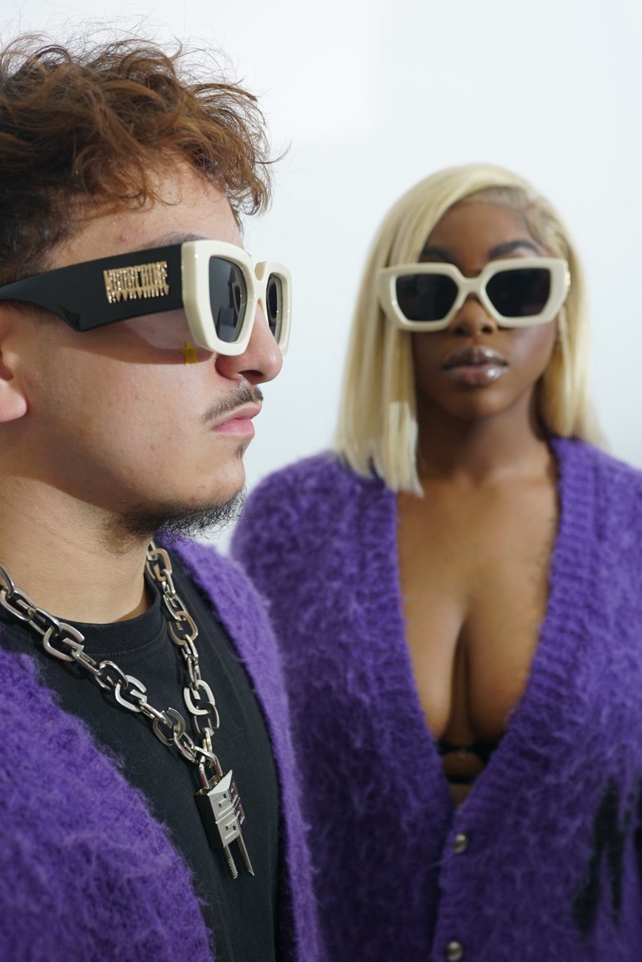 Two models in purple cardigans and cream sunglasses, one in profile in the foreground